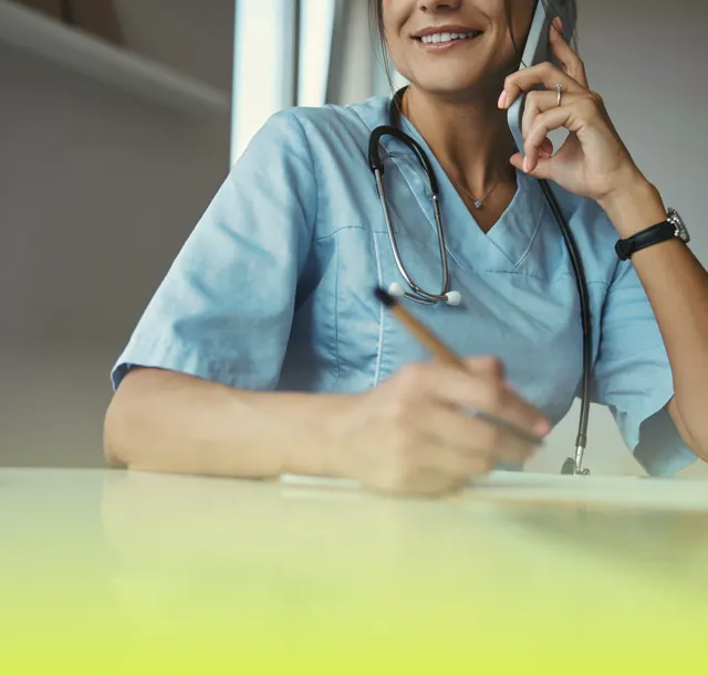 A nurse in blue scrubs smiles as she takes a call at her desk, pen in hand.
